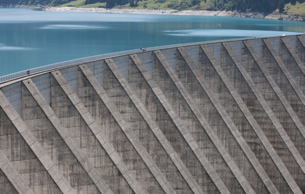 Vue en plongée sur le parement en béton nervuré d'un grand barrage hydroélectrique, avec en arrière-plan un lac aux eaux turquoise et des collines verdoyantes.