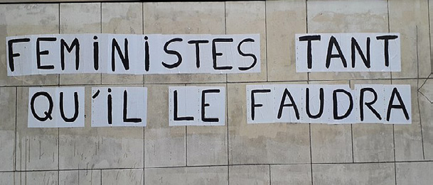 Banderoles militantes de "Colleuses" féministes affichées sur un mur en pierre, portant le slogan « Féministes tant qu'il le faudra ».