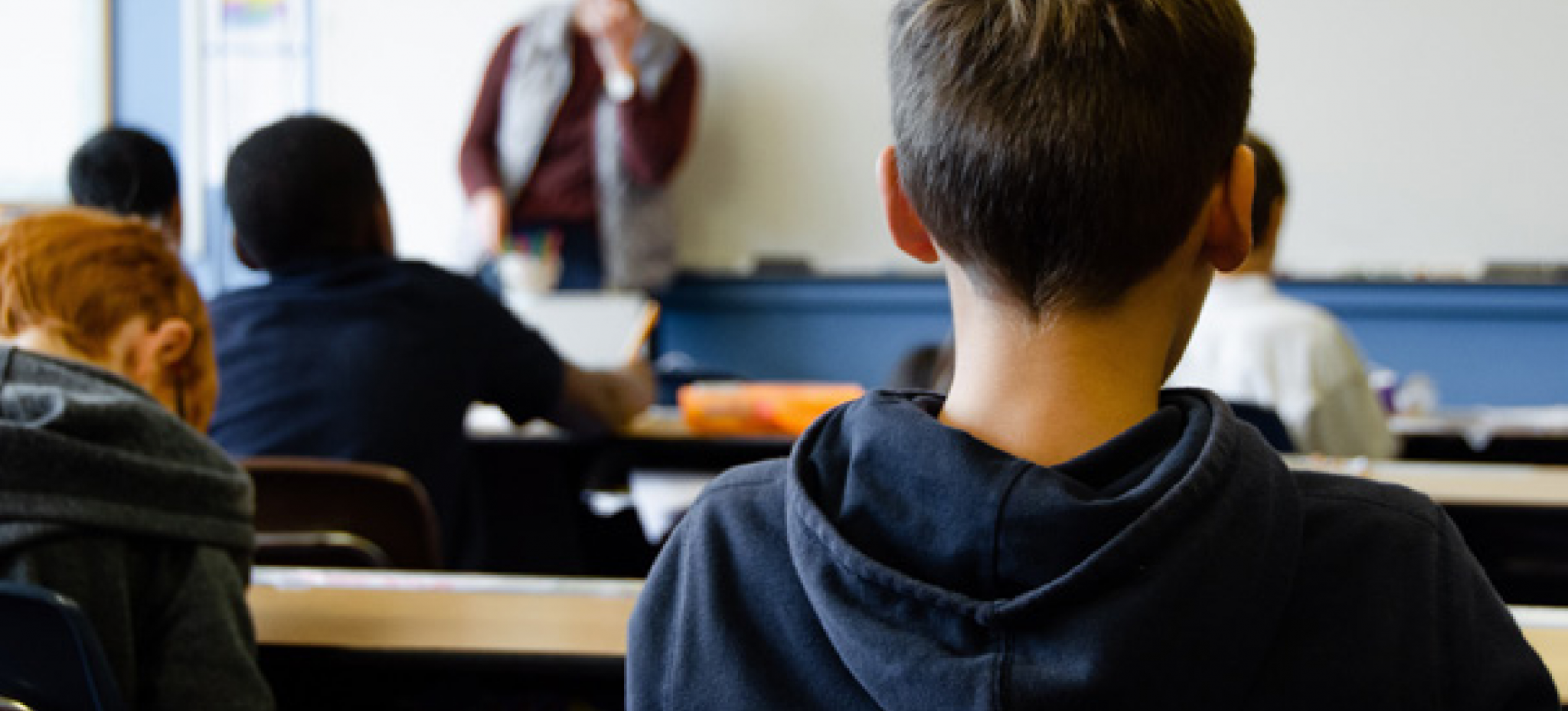 Élèves vus de dos assis à leurs bureaux dans une salle de classe, tournés vers un enseignant debout au tableau en arrière-plan flou.