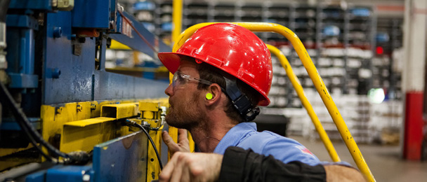 Technicien de maintenance portant un casque de protection rouge, des lunettes de sécurité et des bouchons d'oreilles, effectuant une intervention sur un équipement industriel jaune et bleu dans un atelier de fabrication.