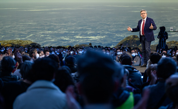 Jean-Luc Mélenchon prenant la parole devant une foule nombreuse réunie en plein air, avec un vaste paysage naturel en arrière-plan.