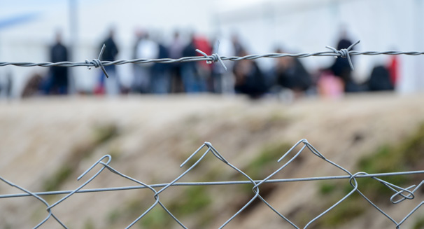 Grillage métallique surmonté de fil barbelé au premier plan, avec un groupe de personnes rassemblées en arrière-plan dans un espace extérieur.