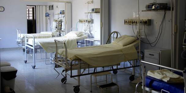 Chambre d'hôpital vide avec plusieurs lits médicaux réglables aux draps beiges, équipée de matériel médical mural et de dispositifs sur roulettes, baignée d'une lumière naturelle tamisée.