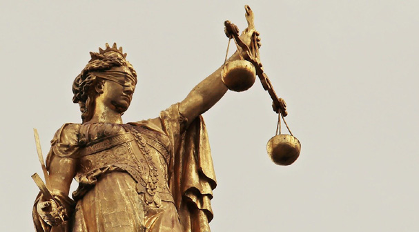 Statue dorée de la Justice les yeux bandés, tenant une balance d'un bras levé et une épée de l'autre, photographiée en contre-plongée sur fond de ciel gris.
