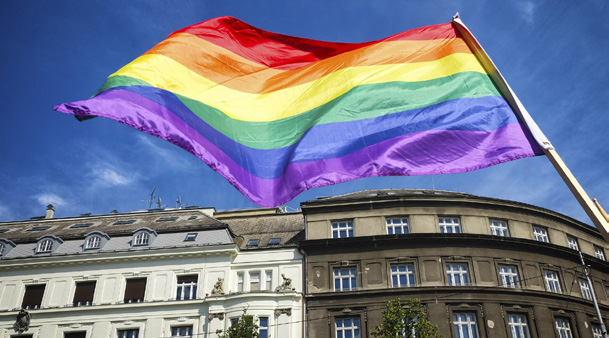 Drapeau arc-en-ciel LGBT+ flottant au vent devant des bâtiments urbains, sous un ciel bleu ensoleillé.