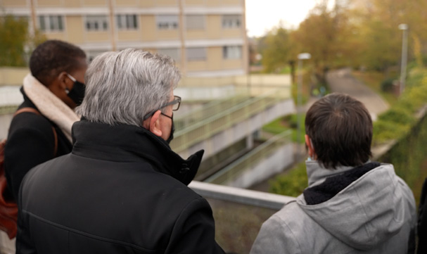 Trois personnes vues de dos (Jean-Luc Mélenchon est au milieu), observent un environnement urbain depuis une passerelle ou un balcon, entouré d'arbres aux feuillages automnaux et d'un bâtiment en arrière-plan.