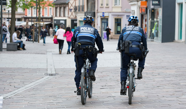 Deux agents de police municipale à vélo, vus de dos, patrouillant dans une rue piétonne commerçante animée.