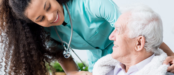 Infirmière en blouse turquoise et stéthoscope souriante, penchée vers un patient âgé aux cheveux blancs, enveloppé dans une couverture, dans un cadre de soin bienveillant.